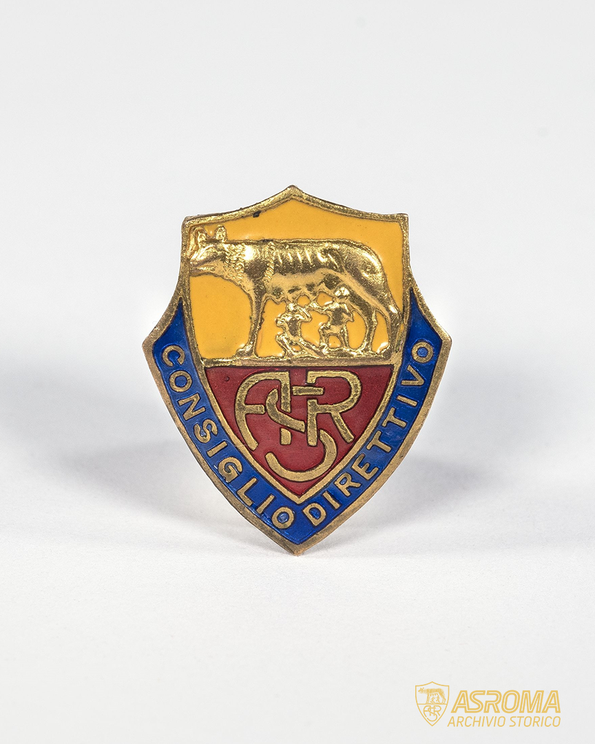 Distintivo del Consiglio Direttivo dell'AS Roma, appartenuto a Italo Foschi. Lo scudo che comprende lo stemma societario è racchiuso dai lati fino al vertice basso da una fascia blu in cui è visibile l'iscrizione "consiglio direttivo". Si tratta, ad oggi, dell'oggetto più antico raffigurante lo stemma originario del Club.
(Archivio Storico AS Roma / Fondo Italo Foschi-Famiglia Zingarelli)